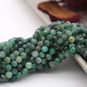Puede incluir: Un hilo de pequeñas cuentas redondas y facetadas de color verde esmeralda. Las cuentas muestran tonos variados de verde bosque profundo y verde menta más claro, con algunas inclusiones oscuras visibles en la superficie de las cuentas.
