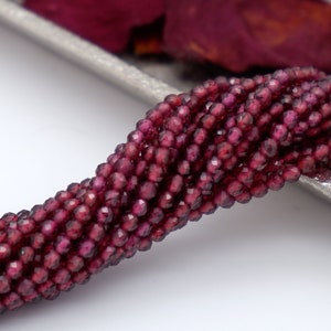 Peut inclure: Gros plan sur des brins de perles de grenat rouge foncé à facettes. Les petites perles rondes ont de multiples facettes qui réfléchissent la lumière. Les perles sont enfilées ensemble, créant une ligne texturée. Le fond est clair, avec un soupçon de plateau argenté.