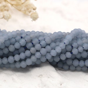May include: Close-up of strands of pale blue faceted beads. The small, round beads are strung together on a dark blue thread. The background is a soft white surface with a hint of dried flowers.