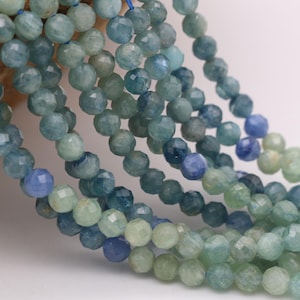 Op de afbeelding: Meerdere strengen gefacetteerde ronde kralen in verschillende tinten blauw, groenblauw en lichtgroen. De kralen hebben een gepolijst, reflecterend oppervlak en zijn geregen aan een dunne blauwe draad, wat zorgt voor een trapsgewijze opstelling.