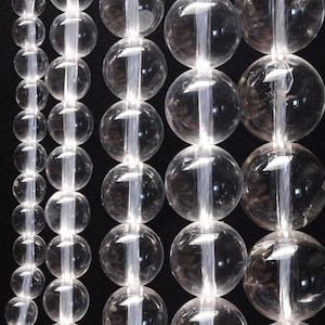 Pode incluir: Um fio de contas de cristal de quartzo transparente, cada conta tem uma forma redonda e lisa. As contas são enfiadas em um fio transparente e estão dispostas em uma linha vertical.
