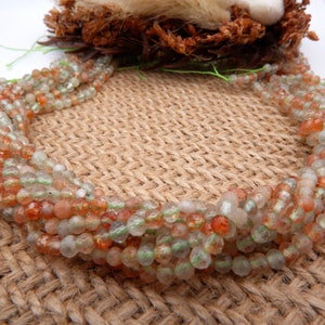 Op de afbeelding: Een streng zonnesteen kralen in tinten oranje, groen en wit. De kralen zijn gefacetteerd en hebben een glinsterende, iriserende afwerking.