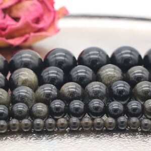 May include: A collection of round, black and gold beads with a shimmering, iridescent effect. The beads are arranged in rows, with the largest beads at the top and the smallest beads at the bottom.