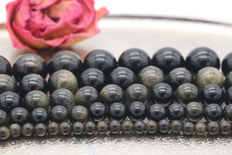 May include: A collection of round, black and gold beads with a shimmering, iridescent effect. The beads are arranged in rows, with the largest beads at the top and the smallest beads at the bottom.