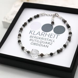 May include: A silver bracelet with clear, black, and gray beads. The bracelet is in a black box with a white label that reads "Klarheit Bergkristall Rutilquarz Obsidian".