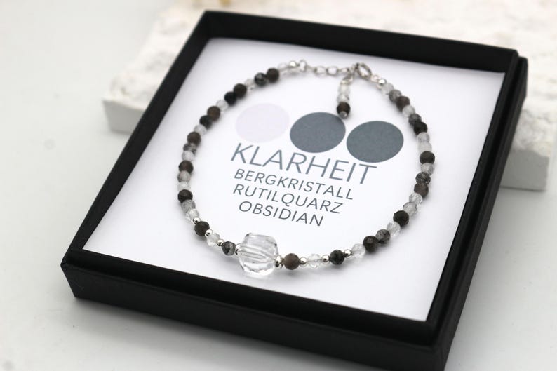 May include: A silver bracelet with clear, black, and gray beads. The bracelet is in a black box with a white label that reads "Klarheit Bergkristall Rutilquarz Obsidian".