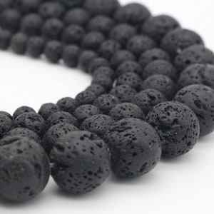 May include: Close-up of a strand of black lava rock beads. The beads are round and have a porous texture. The beads vary in size, with the largest beads in the foreground. The background is white.