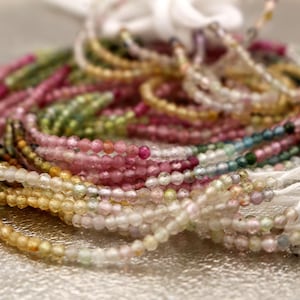 Puede incluir: Una colección de cuentas de piedras preciosas de turmalina multicolor ensartadas en hilo blanco. Las pequeñas cuentas redondas y facetadas muestran un degradado de colores que incluye tonos rosas, verdes, amarillos y transparentes, dispuestas en varios hilos circulares.