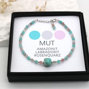 Puede incluir: Un delicado brazalete hecho con cuentas de amazonita, labradorita y cuarzo rosa. El brazalete se muestra en una caja negra con una tarjeta blanca que dice "MUT AMAZONIT LABRADORIT ROSENQUARTZ".