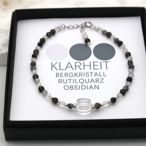 May include: A silver bracelet with clear, black, and gray beads. The bracelet has a large clear crystal charm and the text "KLARHEIT BERGKRISTALL RUTILQUARTZ OBSIDIAN".