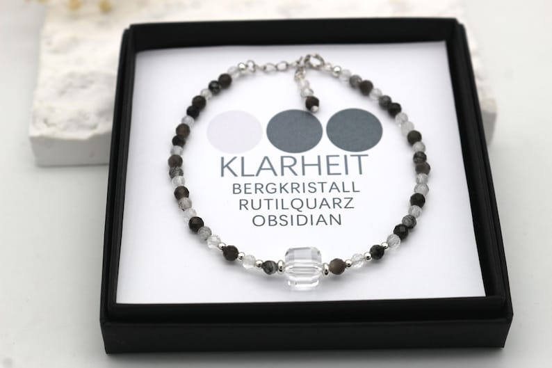 May include: A silver bracelet with clear, black, and gray beads. The bracelet has a large clear crystal charm and the text "KLARHEIT BERGKRISTALL RUTILQUARTZ OBSIDIAN".