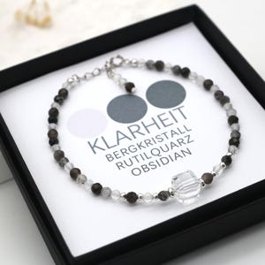 May include: A silver bracelet with black, white, and gray beads. The bracelet has a large clear crystal charm and the text "Klarheit Bergkristall Rutilquarz Obsidian".