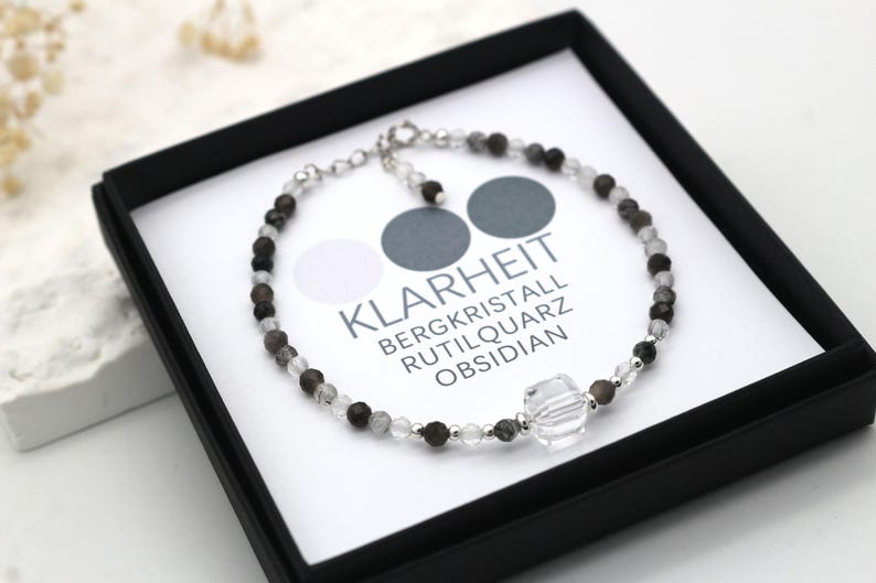 May include: A silver bracelet with black, white, and gray beads. The bracelet has a large clear crystal charm and the text "Klarheit Bergkristall Rutilquarz Obsidian".