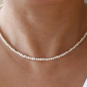 Op de afbeelding: Een eenvoudige witte parelketting. De ketting is gemaakt van kleine, ronde parels die aan elkaar geregen zijn op een dunne ketting. De ketting is delicaat en elegant.