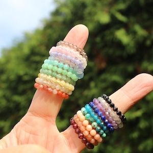 Könnte beinhalten: Ein Stapel bunter Edelsteinringe an einem Finger. Die Ringe bestehen aus verschiedenen Steinen, darunter Amethyst, Rosenquarz und Türkis.