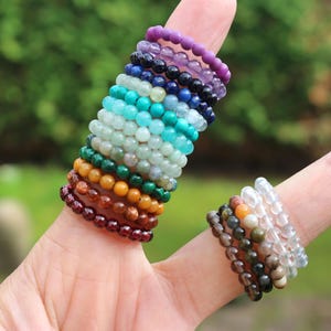 Peut inclure: Une main portant une pile de bagues en pierres précieuses colorées. Les bagues sont faites de petites perles rondes dans différentes nuances de violet, bleu, vert, turquoise, jaune, orange et rouge. Les bagues sont empilées sur l'index, tandis qu'une plus petite pile de bagues est sur le majeur. Les bagues sont faites de perles claires, blanches et brunes.
