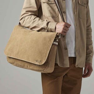 May include: A tan canvas messenger bag with a flap closure and white stitching. The bag has a brown and white striped shoulder strap and the initials "SJ" are visible on the front. The bag is carried over the shoulder.