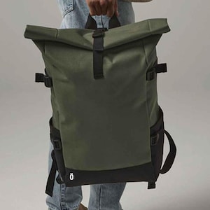 May include: A green and black roll-top backpack with adjustable straps and buckles. The backpack is held by a person wearing blue jeans and tan shoes. The bag has a black base and side pockets.