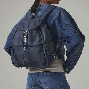 May include: A navy blue backpack with a flap closure, side pockets, and a drawstring. The backpack has a silver-coloured clasp and a small zip pocket. The person in the image is wearing a denim jacket and jeans.