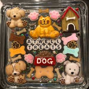 May include: A glass square box with a glittery background and various dog-themed decorations. The decorations include a dog house, dog bones, paw prints, and the words "ATWELL TREATS DOG".