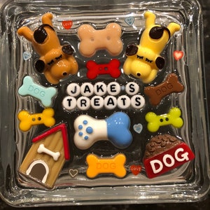 May include: A glass block with a dog-themed design. It features a dog house, bone-shaped decorations, and the words "Jake's Treats" in white letters. There are also several small hearts and the words "Dog", "Love" and "Dog" in various colors.