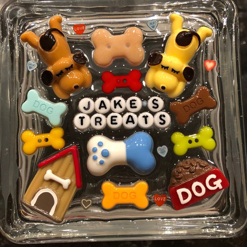 May include: A glass block with a dog-themed design. It features a dog house, bone-shaped decorations, and the words "Jake's Treats" in white letters. There are also several small hearts and the words "Dog", "Love" and "Dog" in various colors.