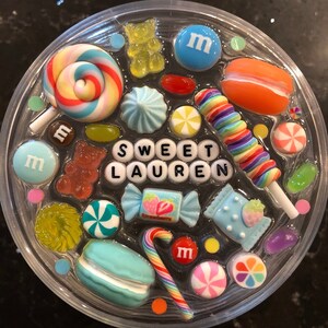 May include: A clear round resin tray with a variety of colorful candy and sweets, including lollipops, gummy bears, macarons, and candy canes. The tray also features the name "SWEET LAUREN" spelled out in white letters.
