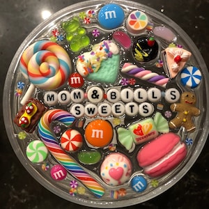 May include: A clear round tray filled with colorful candies, lollipops, and other sweets. The tray has the words "MOM & BILL'S SWEETS" spelled out in white letters.