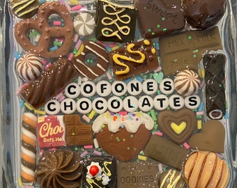 May include: A square glass tray filled with various chocolate candies and treats. The tray is decorated with colorful sprinkles and the words "COFONES CHOCOLATES" are spelled out in white letters. Some of the chocolates include a pretzel, a chocolate bar, a chocolate heart, and a chocolate cookie.