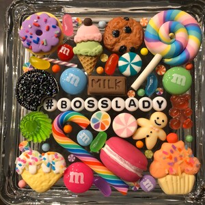 May include: A glass square box filled with colorful candy and sweets, including a donut, ice cream cone, cookie, lollipops, candy canes, gum, and M&Ms. The box also includes a gingerbread man, a cupcake, and a macaron. The text '#BOSS LADY' is spelled out in white letters on the bottom of the box.