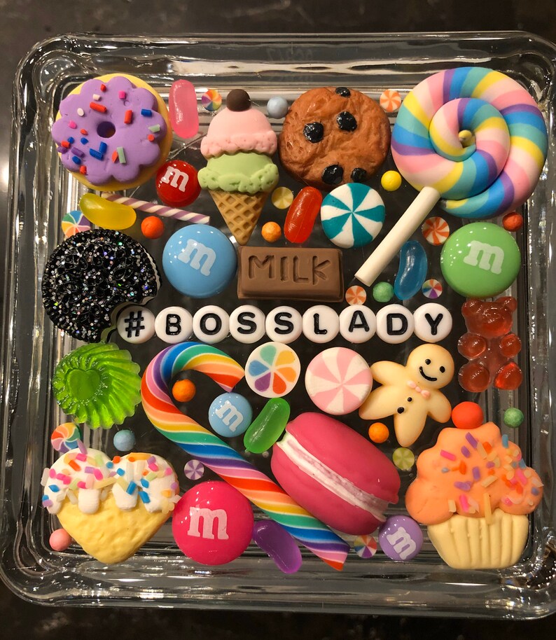 May include: A glass square box filled with colorful candy and sweets, including a donut, ice cream cone, cookie, lollipops, candy canes, gum, and M&Ms. The box also includes a gingerbread man, a cupcake, and a macaron. The text '#BOSS LADY' is spelled out in white letters on the bottom of the box.