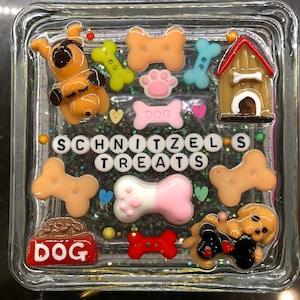 May include: A clear glass square box filled with colorful resin and glitter. The resin includes a dog bone, a dog house, a dog paw print, and a dog bone with a pink paw print. The box also includes a dog bone with the word "DOG" on it, and a set of letter tiles that spell out "SCHNITZEL'S TREATS".