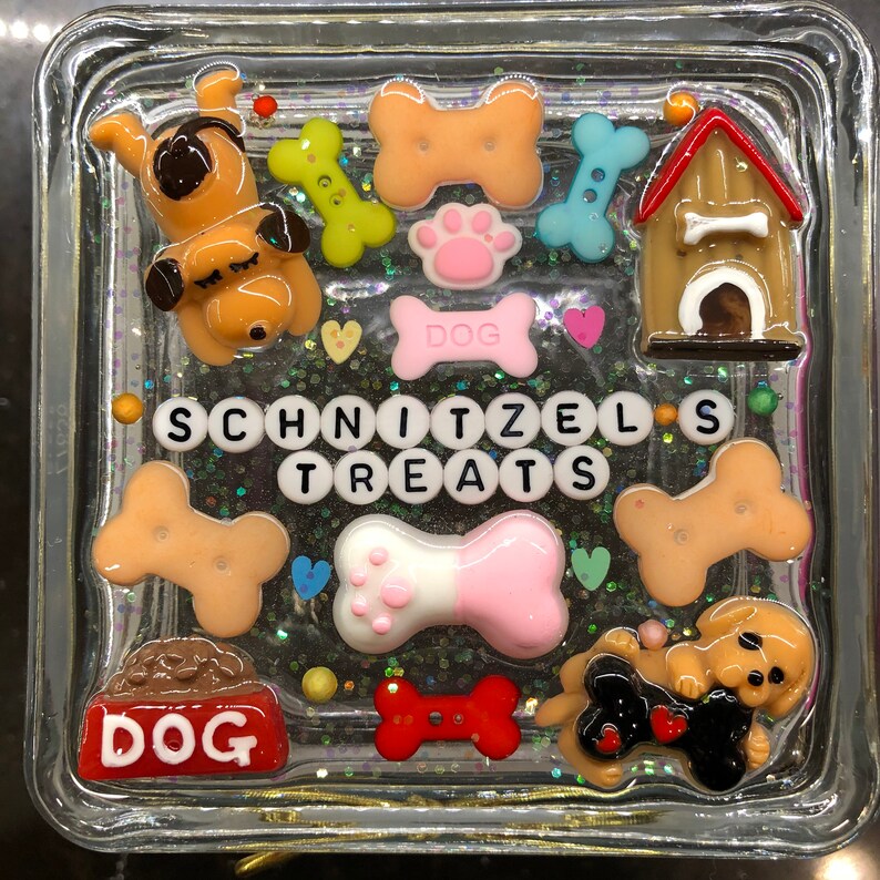 May include: A clear glass square box filled with colorful resin and glitter. The resin includes a dog bone, a dog house, a dog paw print, and a dog bone with a pink paw print. The box also includes a dog bone with the word "DOG" on it, and a set of letter tiles that spell out "SCHNITZEL'S TREATS".
