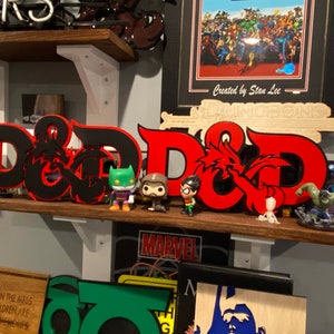 May include: A red and black wooden D&D logo with a dragon design. The logo is on a shelf with other figurines and a framed picture of a comic book cover.