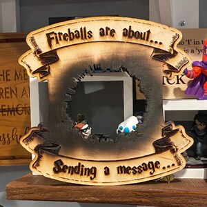 May include: A wooden sign with a burnt-out centre, shaped like a frame. The sign has a banner at the top that reads "Fireballs are about..." and another banner at the bottom that reads "Sending a message..."