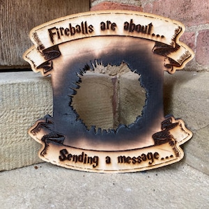 May include: A wooden sign with a burnt-out centre, featuring the text "Fireballs are about... Sending a message..." in a decorative font.