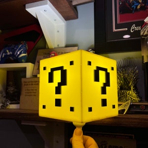 May include: A yellow light-up question block from the Super Mario Bros. video game. The block is made of plastic and has a pixelated design. The block is lit up from the inside, creating a warm glow.