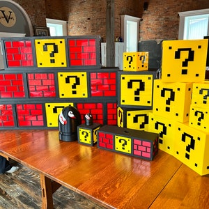 May include: A set of colorful, 3D printed, cardboard blocks featuring the iconic question mark block from the Super Mario Bros. video game. The blocks are stacked in various configurations and are painted in a yellow, black, and red color scheme.