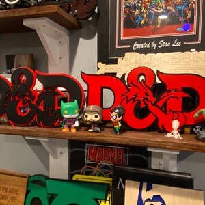 May include: A black and red 3D printed Dungeons & Dragons logo with a dragon design. The logo is on a shelf with several figurines, including a green and black Batman figurine, a brown and black Robin figurine, and a green Hulk figurine.