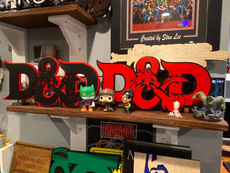 May include: A black and red 3D printed Dungeons & Dragons logo with a dragon design. The logo is on a shelf with several figurines, including a green and black Batman figurine, a brown and black Robin figurine, and a green Hulk figurine.