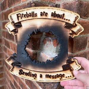 May include: A wooden sign with a burnt-out centre, resembling a fireball. The sign reads "Fireballs are about... Sending a message..."