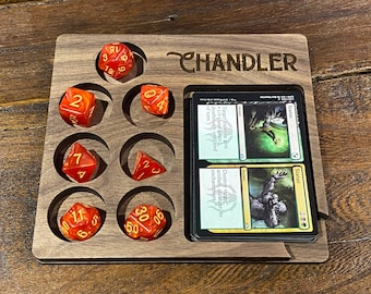 May include: A wooden dice tray with nine compartments for dice and a space for two cards. The tray is engraved with the word "CHANDLER". The dice are red with gold numbers. The cards are Magic: The Gathering cards, one is a green card with the word "Status" and the other is a yellow card with the word "Statue".
