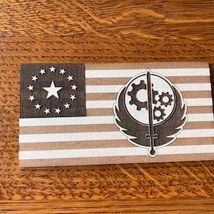 May include: A wooden flag with a brown background and white stripes. The flag features a circle of 13 white stars in the top left corner and a stylized symbol with gears and wings in the right half of the flag.