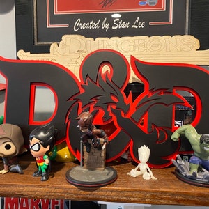 May include: A black and red wooden D&D logo with a dragon design. The logo is surrounded by various figurines, including a green Hulk, a red and green Robin, a black and brown hooded figure, and a small gray figure.