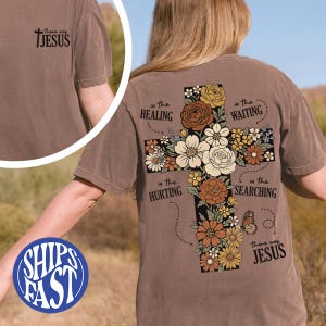 Puede incluir: Camiseta marrón con un diseño de cruz floral. La cruz está adornada con flores blancas, naranjas y amarillas. El texto incluye "in the Healing", "in the Waiting", "in the Hurting", "in the Searching" y "there was Jesus".