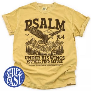Puede incluir: Camiseta de color amarillo mostaza con un gráfico de un águila sobre montañas. El texto dice "PSALM 91:4" y "UNDER HIS WINGS YOU WILL FIND REFUGE". La camiseta tiene mangas enrolladas y un aspecto vintage.