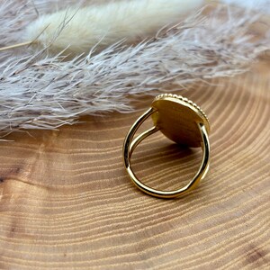K&ouml;nnte beinhalten: Ein goldener Ring mit einer runden, strukturierten Oberfl&auml;che. Das Band des Rings ist schlicht und d&uuml;nn. Die Oberfl&auml;che des Rings ist von kleinen, perlenbesetzten Akzenten umgeben. Der Ring ist vor einem h&ouml;lzernen Hintergrund platziert.