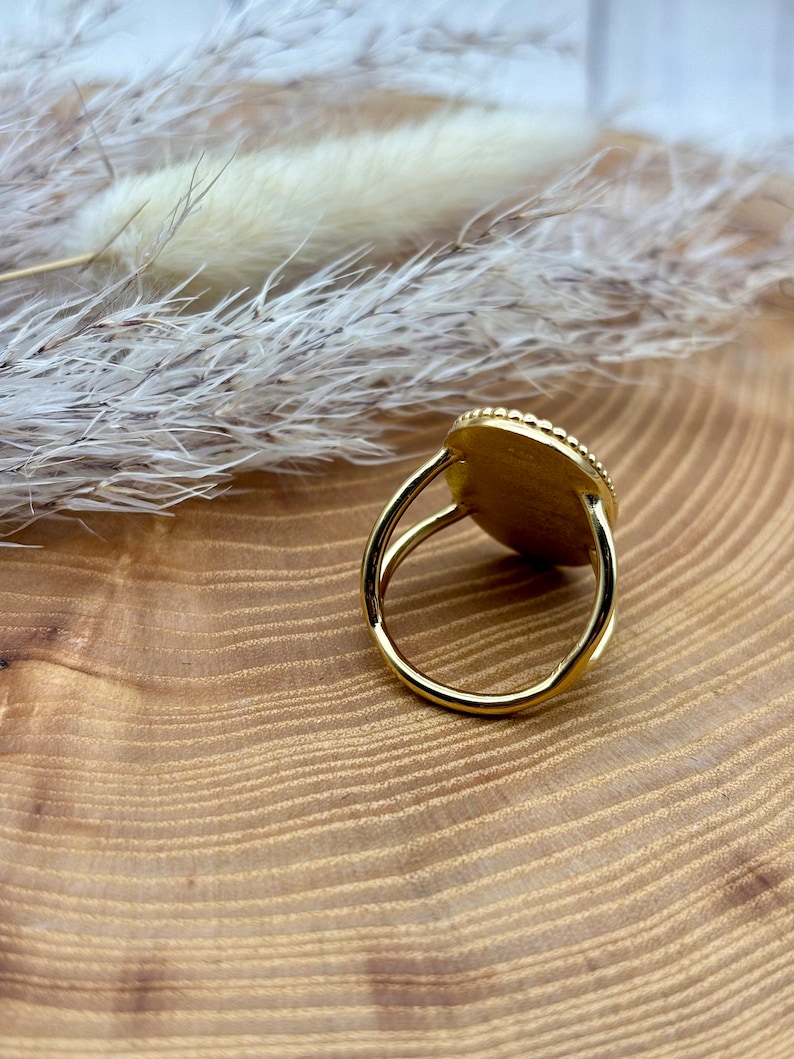 K&ouml;nnte beinhalten: Ein goldener Ring mit einer runden, strukturierten Oberfl&auml;che. Das Band des Rings ist schlicht und d&uuml;nn. Die Oberfl&auml;che des Rings ist von kleinen, perlenbesetzten Akzenten umgeben. Der Ring ist vor einem h&ouml;lzernen Hintergrund platziert.