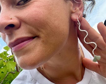 Puede incluir: Pendientes plateados ondulados con un acabado texturizado y un extremo en forma de hoja. Los pendientes son largos y colgantes, creando un aspecto único y elegante. La joyería se usa con una camiseta blanca y un delicado collar de oro.
