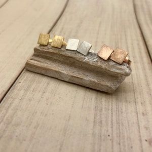 Könnte beinhalten: Fünf Paar quadratische Ohrringe in verschiedenen Metalloberflächen: Gold, Silber und Roségold. Die Ohrringe werden auf einem Stück Treibholz präsentiert.